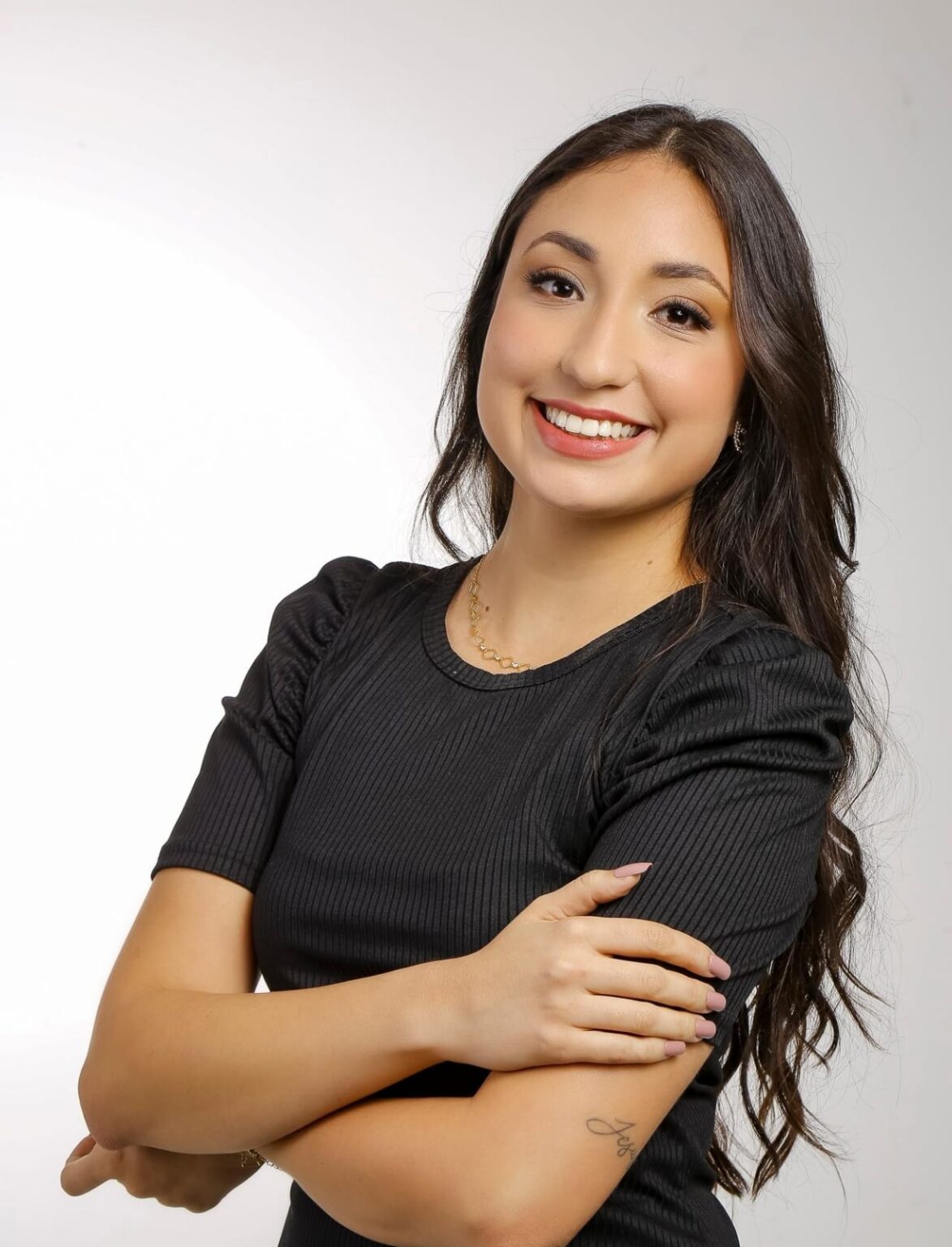 jovem mulher sorrindo com fundo claro neutro, posando para uma fotografia profissional com os braços cruzados